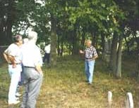 a mature man wearing jeans and plaid shirt presents a daytime lecture to several other persons while all stand amongst old tombstones and trees.