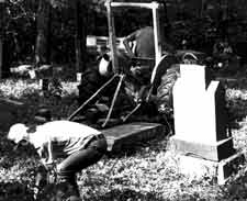 a man clears brush and a small tractor lifts a fallen headstone during a cemetery restoration work day.