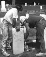 two individuals in casual work clothes install new white stone veterans grave markers during a restoration in an historic city cemetery.