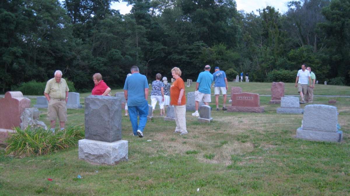 a dozen people in summer clothing standing and walking amongst grey and red granite grave markers in a flat green grass cemetery park.