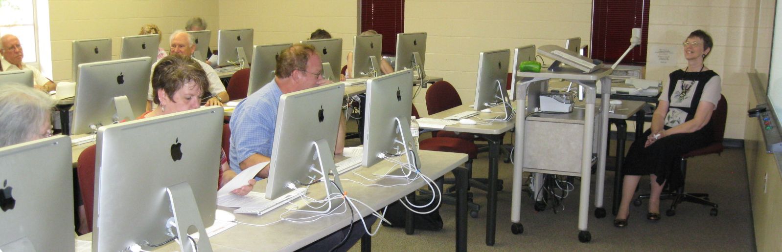 rows of tables with Apple brand desktop computers, attendees of online workshop, overhead projector and white board at front of classroom