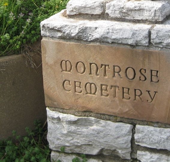 three dimensional structure built of gray flagstone inset with brown sandstone engraved with cemetery name, green plants grow close by.